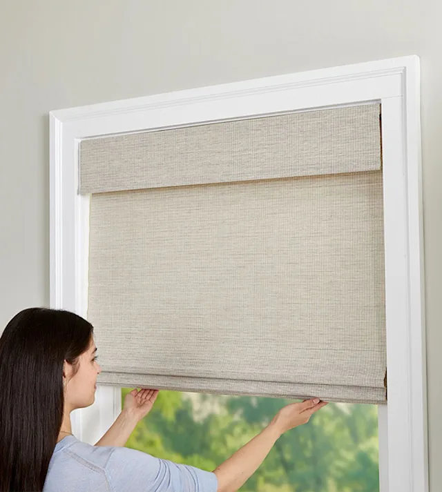 A lady demonstrating a cordless lift natural woven shade, lifting it with a light push, in a Nicholasville, KY showroom with sleek wooden accents and soft spotlighting.