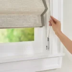 Natural woven shade with a continuous loop bead chain system, featuring a child-safe tensioner, showcased on a display in a Nicholasville, KY showroom, framed by sleek wooden trim and soft spotlighting.