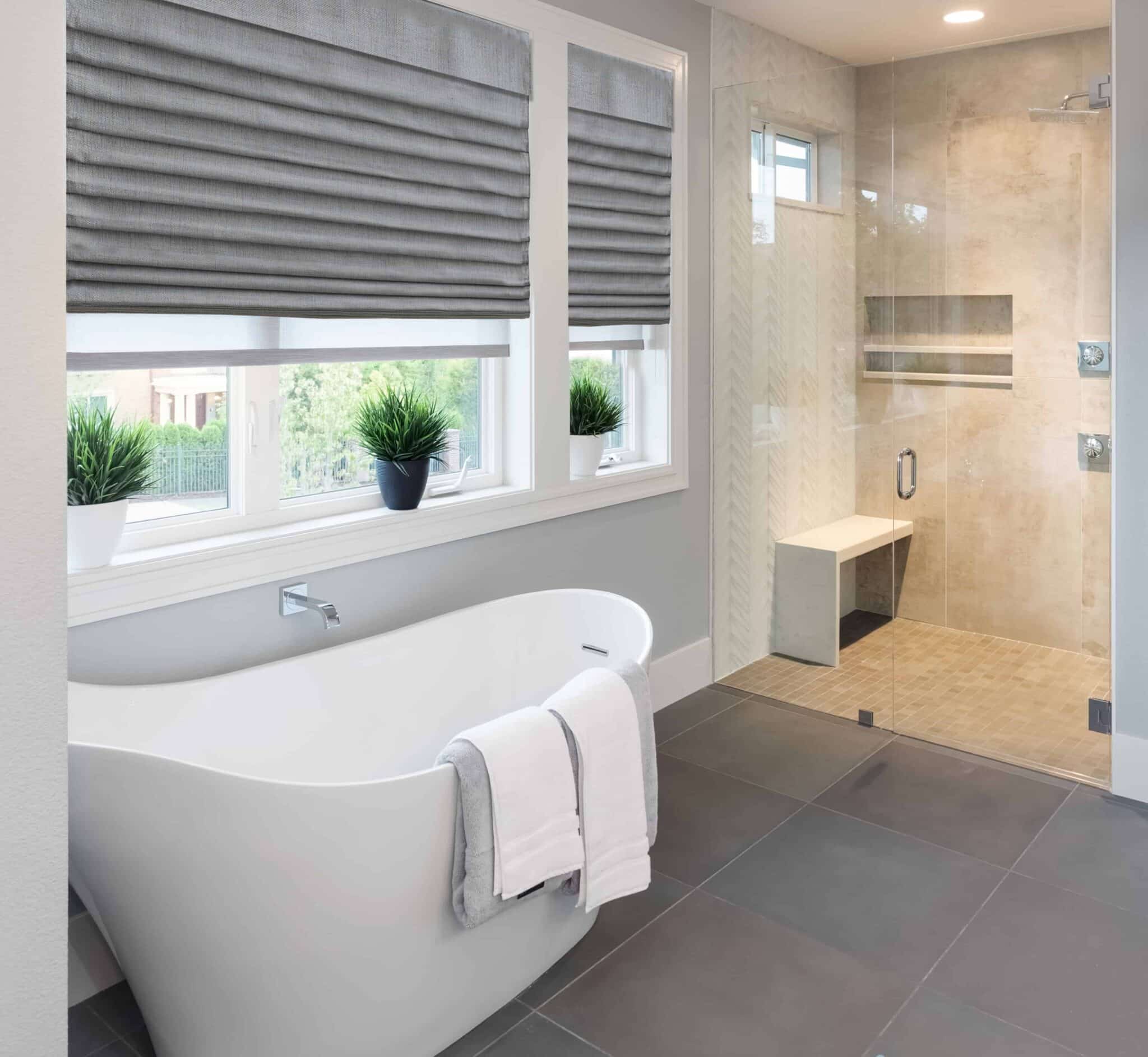 Window Shades 10 Flat Roman shades cover a double window in a bathroom, softly filtering light. A bathtub is positioned in front of the window, creating a serene and private atmosphere with elegant decor.