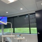Charcoal-colored roller shades are installed on the windows of a treatment room at Dental Concepts and Implant Center in Lexington, KY. The shades softly filter natural light, creating a modern and welcoming atmosphere in the dental office, which features a dental chair and advanced technology