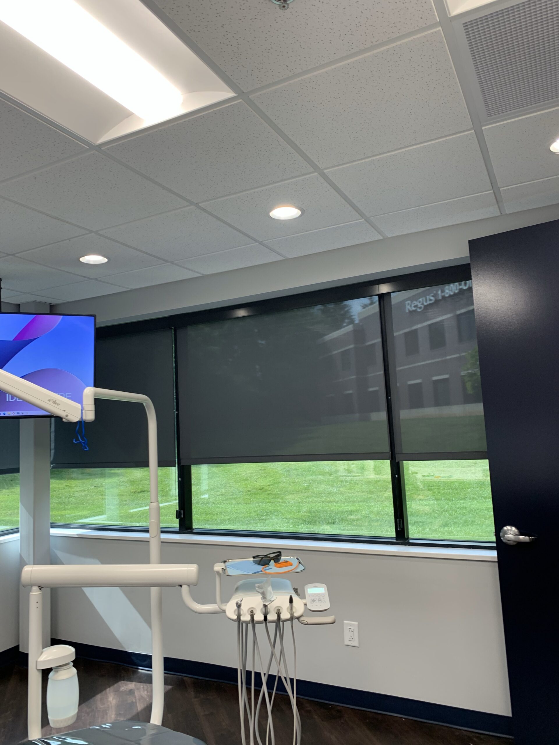 Charcoal-colored roller shades are installed on the windows of a treatment room at Dental Concepts and Implant Center in Lexington, KY. The shades softly filter natural light, creating a modern and welcoming atmosphere in the dental office, which features a dental chair and advanced technology